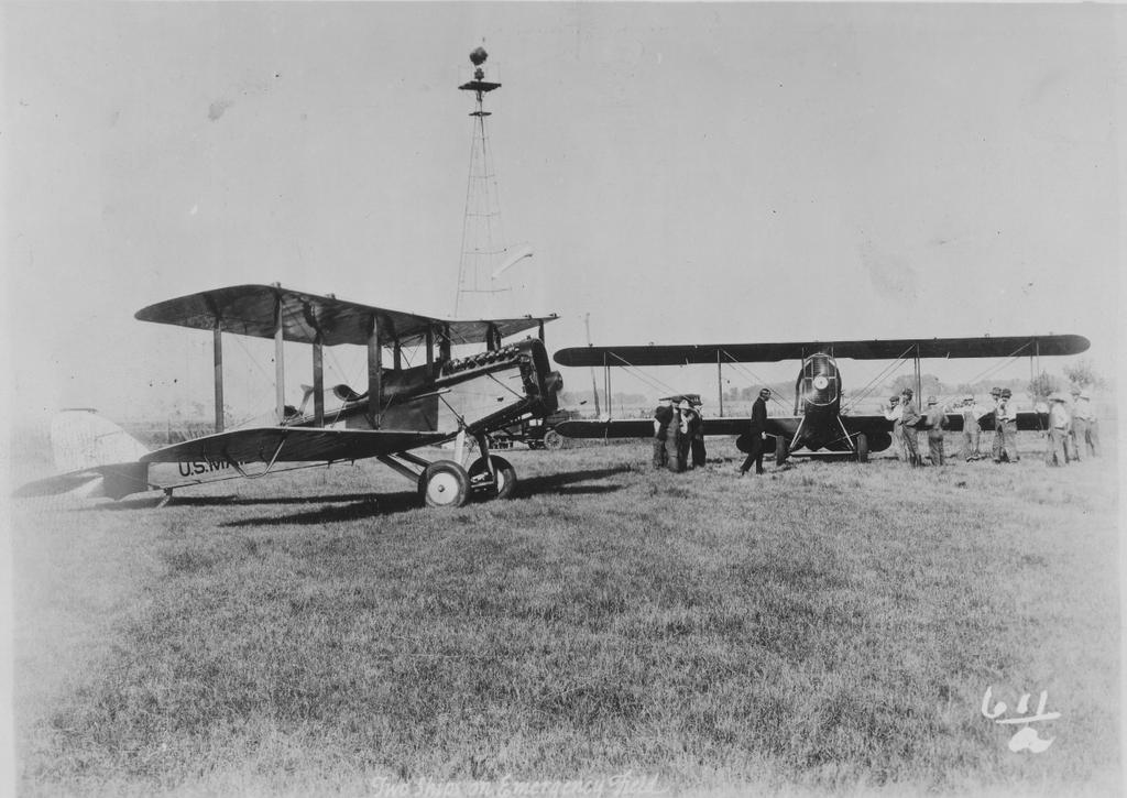 Airmail planes, pilots, and onlookers at unidentified airfield, circa 1924. In the background stands a beacon light tower, which helped guide pilots at night.