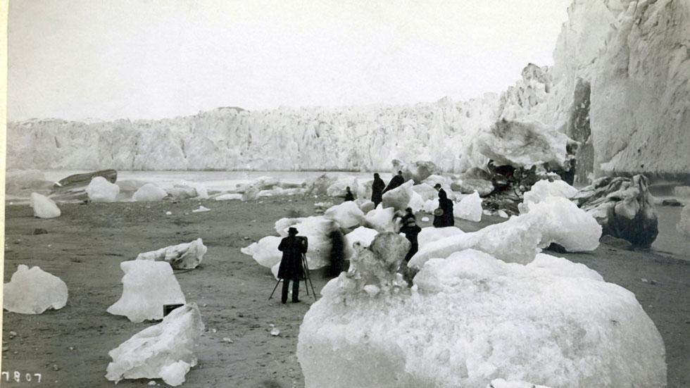 A photographer and several tourists explore the icebergs in Muir Inlet sometime in the 1880s or 1890s, while in the background the glacier rises more